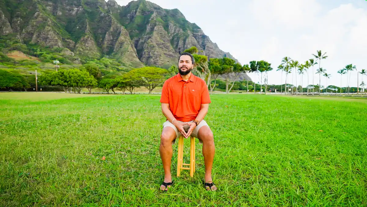 Daniel Molina sitting on a stool in a field below the Ko'olau mountains on O'ahu
